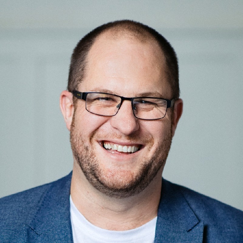 A picture of Ant smiling. He has brown hair, a slight beard, wears glasses. He is wearing a blue jacket with a white t-shirt.